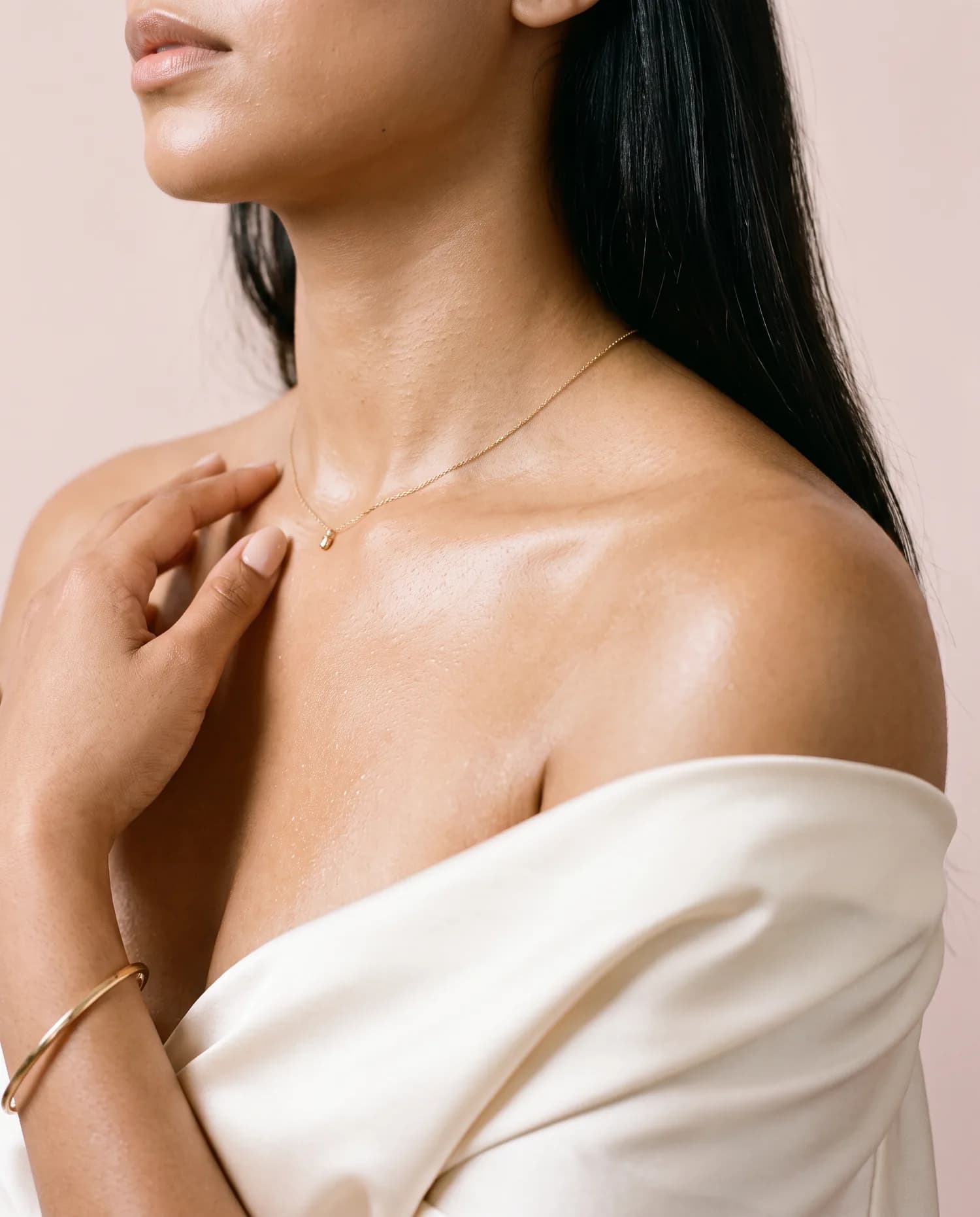 Golden body oil glistening on a woman's collarbone above an ivory satin drape