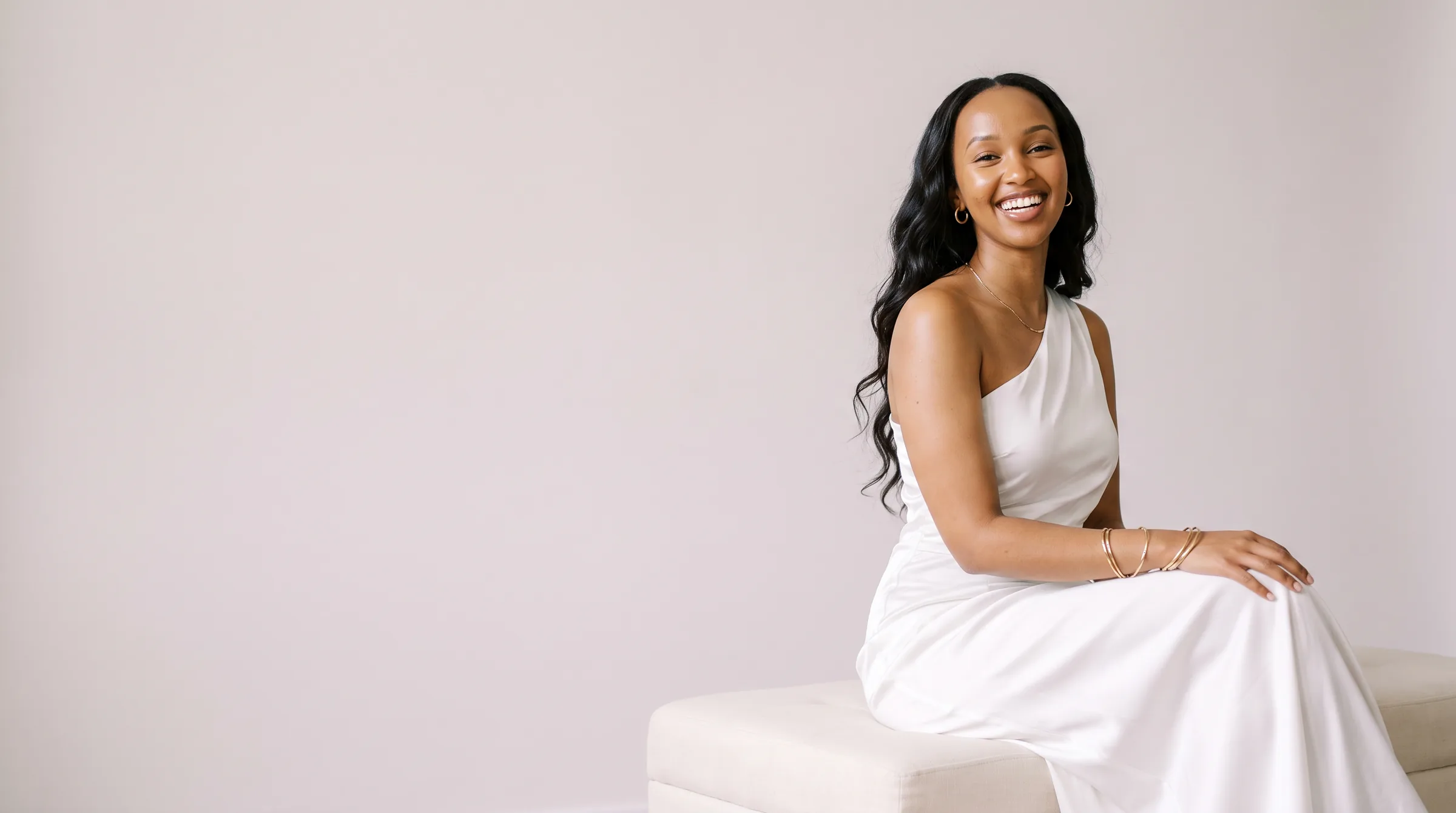 Campaign image: a laughing young woman with glowing light skin and long dark waves sits on an ivory bench in a white satin gown and gold bangles